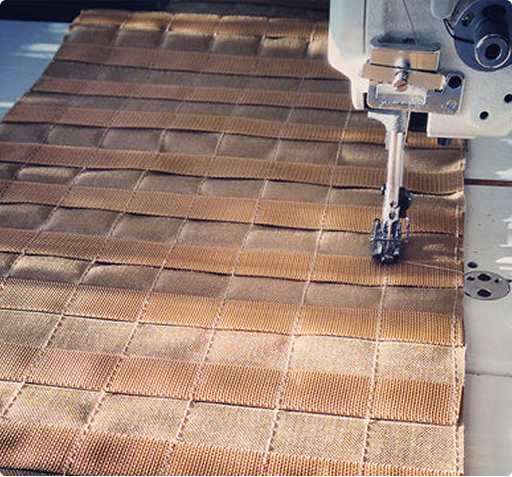 A close-up of an industrial sewing machine stitching a series of woven fabric strips, demonstrating precision manufacturing in tactical gear production